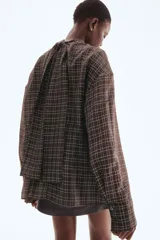 Blusa de manga larga con estampado a cuadros en tonos oscuros (negro/marrón y blanco). Presenta cuello drapeado con tira para atar en la nuca, hombros caídos y cintura entallada con pinzas moldeadoras.