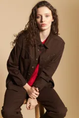 Sobrecamisa de gabardina color marrón, con cierre frontal de botones, bolsillos en el pecho y detalle de tachas metálicas en los hombros y canesú. Presenta cuello clásico y mangas largas con puños abotonados.