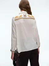 Camisa de color crudo con detalles de bordados en tonos mostaza y marrón en el cuello, hombros, bolsillos frontales y puños. Presenta cierre frontal con botones y dos bolsillos tipo cargo en el pecho.