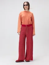 Pantalón de punto jacquard con diseño geométrico en tonos rojos. Presenta un corte holgado de pierna ancha, tiro medio, cintura elástica con cordón ajustable y una franja lateral lisa en contraste.