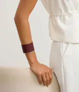 Pulsera rígida de metal con acabado en poliuretano color bordó y detalles dorados. Presenta una abertura para facilitar su colocación.