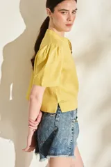 Blusa de lino color amarillo, con cuello mao, cierre frontal con cremallera y mangas cortas abullonadas. Cuenta con un bolsillo aplicado en el frente.