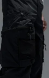 Pantalón cargo oversize de corte relajado, confeccionado en textil técnico resistente de color negro. Presenta bolsillos laterales cargo de gran tamaño con cierres metálicos, bolsillos laterales superiores y bolsillos traseros con broches a presión. Incluye cinturón integrado de cinta de seguridad con hebilla de encastre técnica.