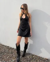 Vestido corto negro sin mangas, con escote halter y detalle de abertura tipo gota en el pecho. Presenta fruncidos laterales en la cintura y falda evasé.