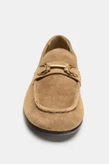 Mocasín de ante color marrón claro, con detalles de pespuntes en relieve, aplique metálico dorado en el empeine y suela plana con pequeño tacón.