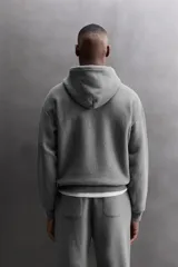 Sudadera hoodie gris de corte relaxed fit, confeccionada en tejido de algodón con interior afelpado. Presenta cuello con capucha ajustable, manga larga, texto estampado en color bordó en el pecho, bolsillo frontal tipo canguro, aplicación de parche con logo en la parte inferior y acabados en rib.