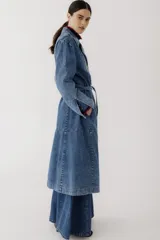 Trench largo de jean azul con doble botonadura, cinturón y bordado floral en el cuello.