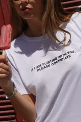 Remera blanca de algodón con cuello redondo y manga corta, con texto estampado en negro en el pecho que dice "If I am flirting with you, please cooperate".