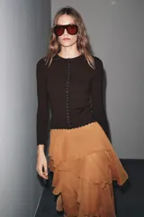 Chaqueta ajustada de punto acanalado color marrón oscuro, con cuello redondo y manga larga. Presenta un cierre frontal completo con botones pequeños y blancos. Los bordes del ruedo y los puños tienen un acabado festoneado.