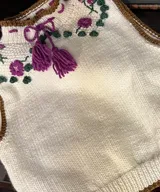 Chaleco tejido color crudo con bordados florales en tonos magenta y verde, cuello alto con cordón y borlas magenta, y terminaciones en marrón.