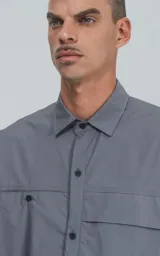 Camisa de manga corta en color gris, confeccionada en popelina de algodón. Presenta un corte oversize, cuello clásico, cierre frontal con botones y dos bolsillos delanteros, uno de ellos con solapa.