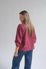 Blusa bordó de corte holgado, con escote en V y cuello fruncido. Presenta mangas tres cuartos con puños elásticos y detalles de volados.