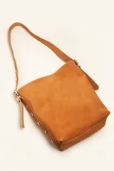 Cartera tipo balde color tostado, confeccionada en gamuza, con correa ajustable decorada con tachas metálicas plateadas.