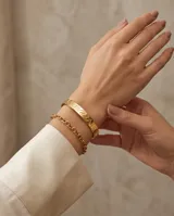 Esclava rígida de acero con baño de oro, con diseño grabado en relieve. Se presenta junto a una pulsera de cadena fina de eslabones.