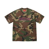 Camiseta de manga corta con estampado de camuflaje en tonos verdes y marrones. Presenta el logo de la marca bordado en el pecho con letras cursivas en color violeta con contorno amarillo.