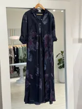 Vestido largo color azul marino con estampado floral en tonos violeta y bordó, estilo camisero con cuello tipo solapa, cierre frontal parcial con botones y mangas largas remangables.