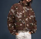 Campera puffer color marrón habano con estampado de loros, estrellas y elementos botánicos, cuello alto y cierre frontal.