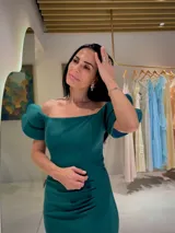 Vestido largo de fiesta color verde esmeralda, con escote recto estilo hombros caídos y mangas cortas con volados estructurados. Presenta un detalle de fruncido lateral en la cintura que aporta un ajuste entallado a la silueta.