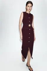 Vestido midi de cuello redondo y sin mangas, confeccionado en tejido tipo crepe. Presenta cierre frontal con botones dorados, bolsillos con solapa en la parte delantera y un cinturón elástico integrado con hebilla metálica decorativa.