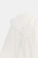 Camisa blanca de manga larga con escote en V y volantes superpuestos en el borde del escote. La tela presenta una textura sutilmente arrugada o jacquard.