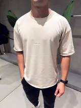 Remera de algodón pesado color beige, con corte oversize, cuello redondo y bordado pequeño en el centro del pecho. Presenta terminaciones con bordes sin costura en mangas y ruedo.