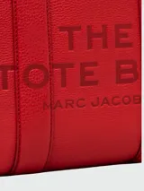 Cartera tipo tote de cuero rojo de la marca Marc Jacobs, modelo The Leather Small Tote Bag. Presenta doble asa superior, correa ajustable y extraíble para el hombro, y el texto grabado en relieve "THE TOTE BAG MARC JACOBS" en el frente.