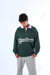 Polo de frisa color verde oscuro con cuello blanco y cierre parcial frontal. Presenta un bordado en el frente con el texto "Green Dreams" en tipografía cursiva blanca y la palabra "Aum" debajo.