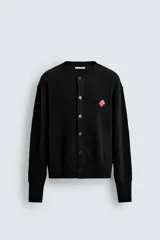 Cárdigan de punto negro, corte relajado, con cuello redondo, manga larga y cierre frontal de botones oscuros. Presenta un parche bordado en forma de dado rojo con corazones blancos en el lado izquierdo del pecho.