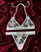 Conjunto de lencería de tul bordado blanco, compuesto por un corpiño triangular con tirantes halter, moños decorativos y la inscripción 'PURO FUEGO' en strass, y una bombacha tipo colaless con tiras cruzadas en el frente y la inscripción 'PARA VOS' en strass.