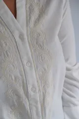Blusa blanca de manga larga con cuello mao y cierre frontal de botones. Presenta detalles de bordados decorativos en los laterales del frente y puños anchos.