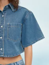 Camisa corta de denim azul claro, con cuello camisero, manga corta ancha y cierre frontal de botones blancos. Presenta un bolsillo de parche en el pecho izquierdo.
