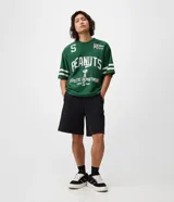Remera de corte boxy y estilo deportivo, color verde oscuro, con diseño inspirado en el fútbol americano de Peanuts. Presenta cuello redondo, mangas cortas con franjas blancas horizontales y estampas localizadas de Snoopy en el frente.