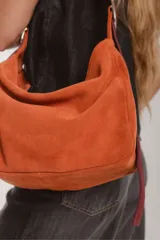 Bolso tipo hobo de gamuza color naranja quemado, con correa ancha de tela color bordó.