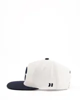 Gorra de béisbol blanca con visera plana azul marino y logo bordado en el frente.