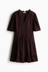 Vestido corto de color bordó, con escote en V y cuello de volados, mangas cortas abullonadas y botonadura frontal con alforzas e inserciones de encaje. Presenta costuras angulares con frunces en la cadera y falda con ribete de volado en el bajo.
