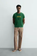 Camiseta de corte relajado, color verde, con cuello redondo y manga corta. Presenta un texto bordado en relieve con letras amarillas y contorno oscuro en el centro del pecho.
