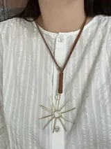 Set de accesorios compuesto por un brazalete rígido abierto, un collar con dije de sol radiante, un par de caravanas onduladas y un par de caravanas tipo fleco, presentados en un sobre de cuero con costuras artesanales.