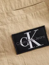 Camisa de manga larga color beige, confeccionada en tejido texturizado de algodón. Presenta cuello camisero, cierre frontal de botones y un bolsillo en el pecho con una insignia cuadrada negra con el logo 'CK' en blanco.