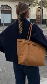 Cartera tipo tote de símil gamuza color marrón, con diseño estructurado y amplio, asas de hombro delgadas y detalle de tira con hebilla y argollas metálicas en la parte superior.