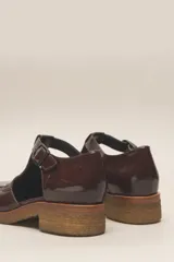 Zapato tipo guillermina de cuero charol color marrón oscuro, con tira T y hebilla ajustable en el lateral. Posee puntera cuadrada y suela de goma color caramelo con taco bajo.
