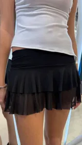 Minifalda negra de tiro bajo con diseño de doble volado, confeccionada con una capa superior de tela opaca y una capa inferior de tul transparente. Incluye short interno incorporado.