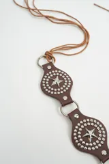 Cinturón negro de estilo western con apliques metálicos circulares y estrellas grabadas, unido por argollas metálicas y terminado con tiras largas de cuero para atar.