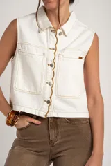 Chaleco de denim blanco con diseño sin mangas, cuello camisero, bolsillos frontales con solapa y cierre de botones metálicos con detalle de ribete ondulado en contraste.