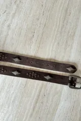 Cinturón de gamuza color chocolate con apliques metálicos en forma de rombo y tachas decorativas. Cuenta con hebilla metálica de estilo rústico.