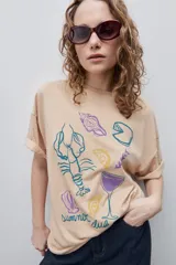 Remera color tostado de corte holgado, con cuello redondo y mangas cortas. Presenta un estampado gráfico frontal de estilo veraniego con ilustraciones de una langosta, conchas, limón, una copa y la inscripción 'Summer Club'.