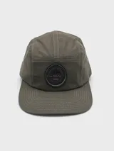 Gorra verde con visera plana y parche circular negro con logo de montaña.