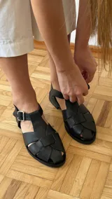 Sandalia de cuero marrón con diseño trenzado artesanal en el empeine, pulsera ajustable al tobillo con hebilla metálica y suela plana.