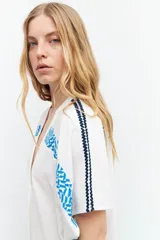 Túnica blanca de lino con corte oversized, escote en V, cierre con botones y mangas cortas. Presenta bordados geométricos azules en hombros y laterales.