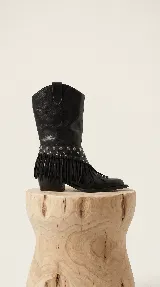 Bota estilo texana de cuero negro, con taco bajo y caña media. Presenta un aplique desmontable en la zona del tobillo decorado con tachas metálicas y flecos largos.