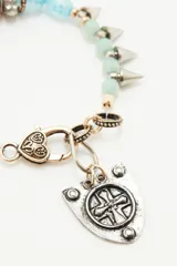 Pulsera de diseño artesanal compuesta por una combinación de cuentas de vidrio en tonos rojizos y negros, intercaladas con piezas metálicas. Presenta un dije central en forma de escudo con detalle de cruz grabada y cierre de mosquetón.